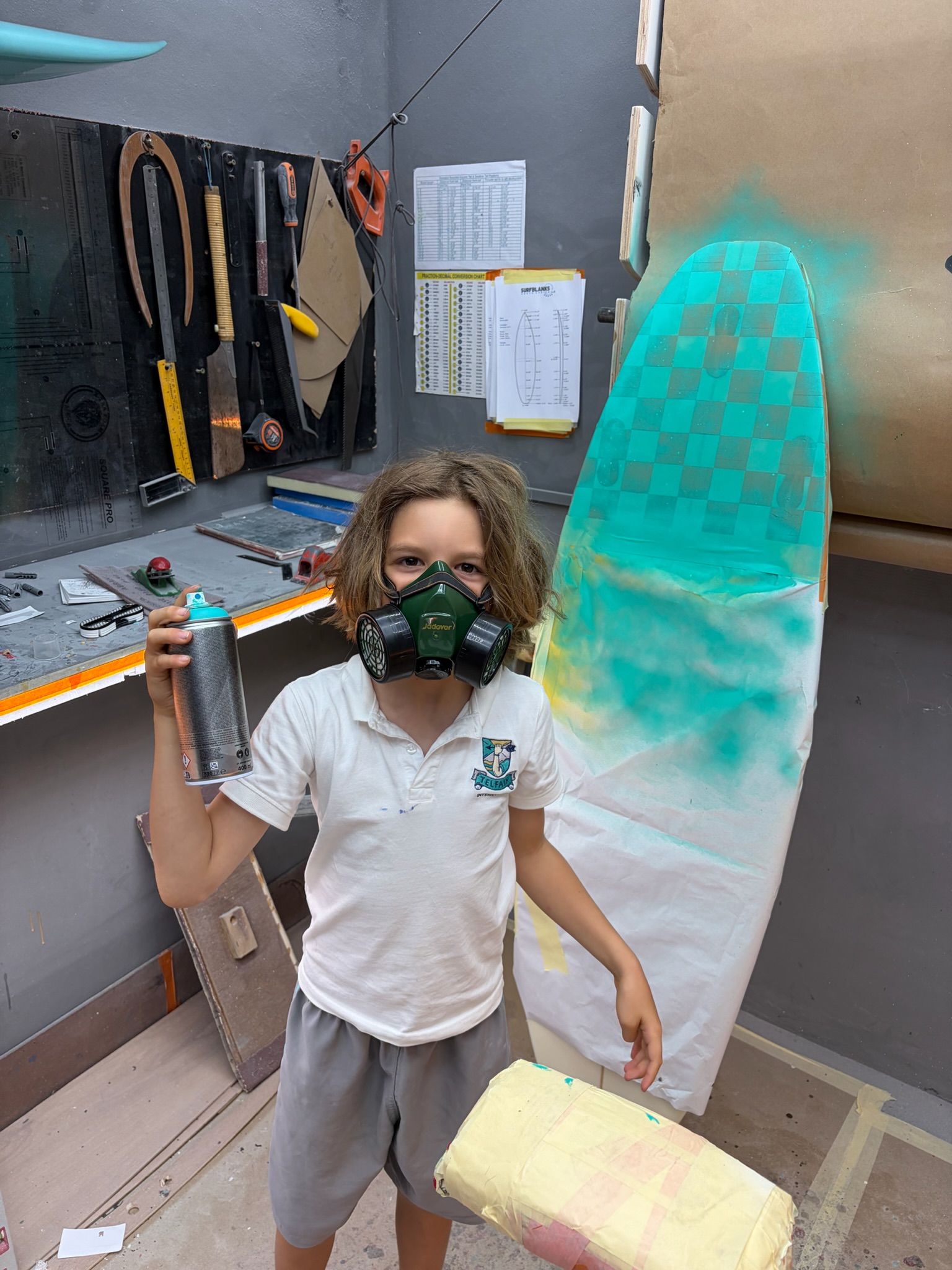 Noah in a green respirator holding a spray can next to the board with its freshly sprayed teal checkerboard tail