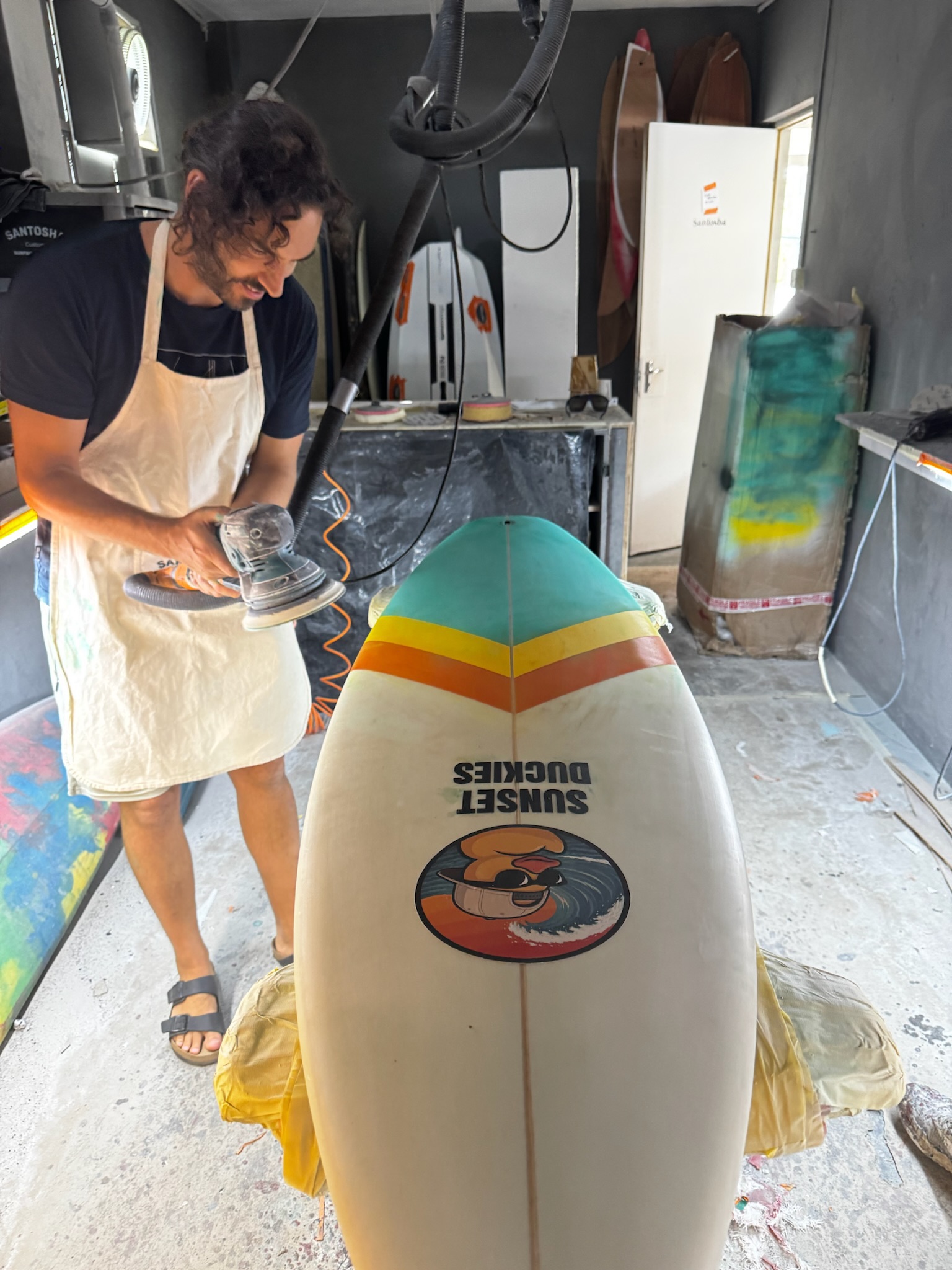 A shaper orbital-sanding the glassed deck, the full Sunset Duckies graphic visible